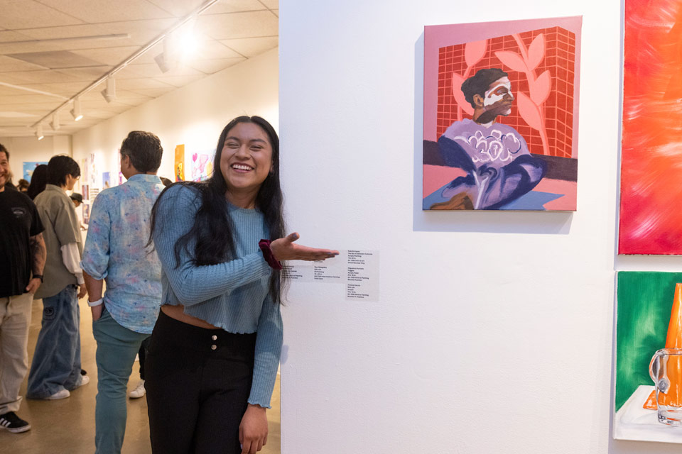 A student stands adjacent to and gestures to their painting displayed on a wall.